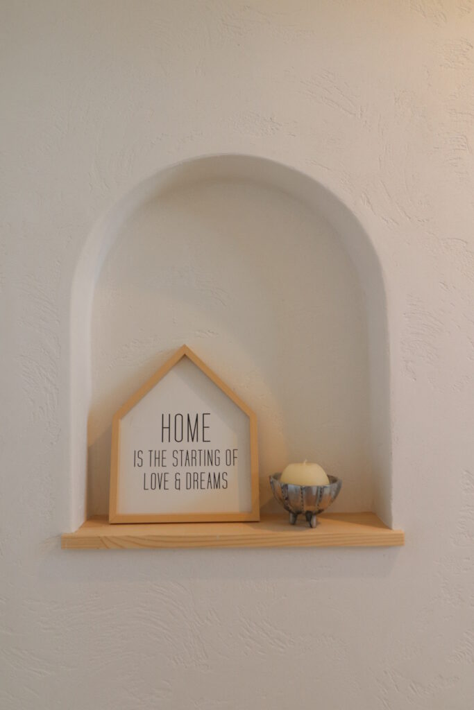白い漆喰風の壁にあるアーチ型のニッチ。「HOME IS THE STARTING OF LOVE & DREAMS」と書かれた家型の木製フレームとキャンドルが飾られたインテリア。|盛岡市で平屋をお考えの方は高断熱高気密注文住宅の大共ホーム
の画像
