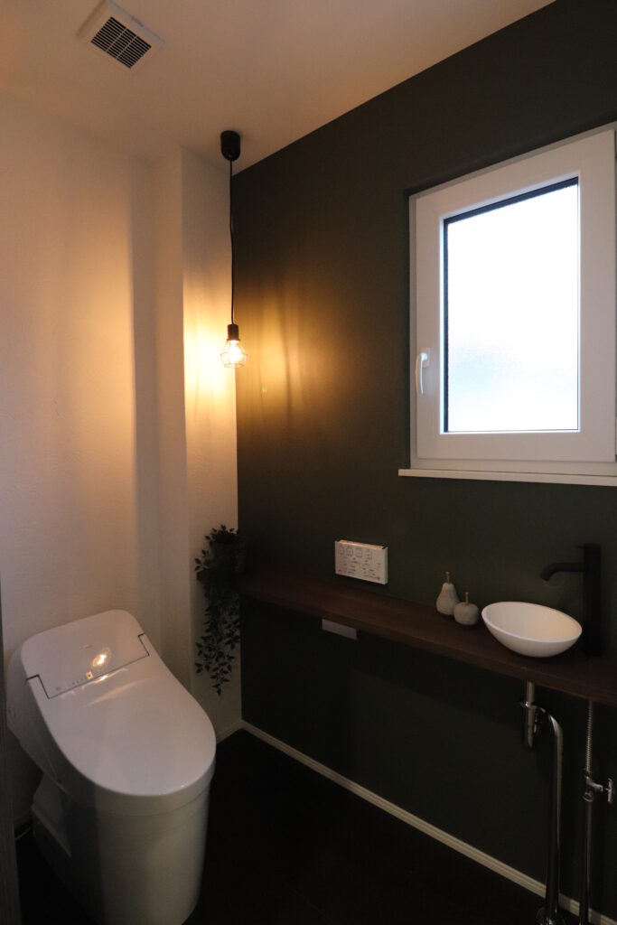 深みのあるダークグリーンのアクセントクロスに裸電球のペンダントライトが灯るシックなトイレの画像