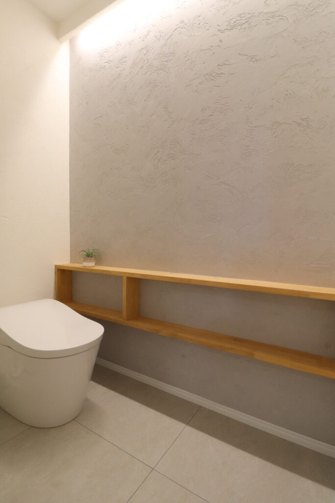 グレーの塗り壁に木製のオープン造作棚と上部間接照明を施したモダンなトイレの画像