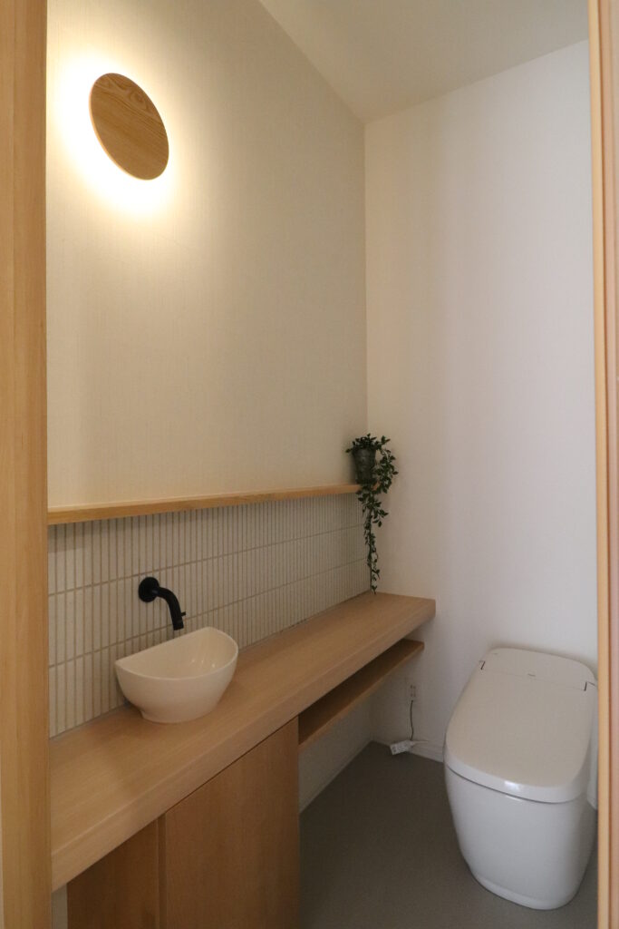 明るい木目調カウンターに縦長ボーダータイルと丸型ブラケットライトを合わせた和モダンなトイレの画像