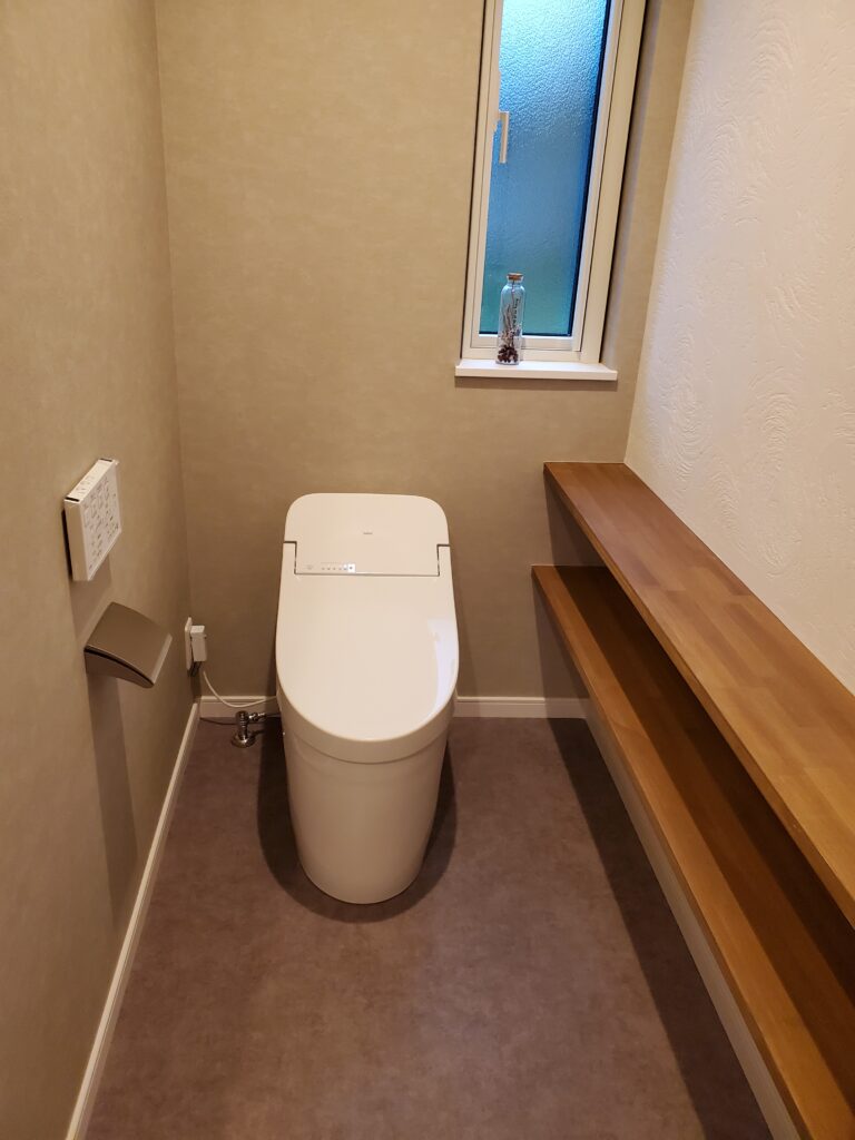 ベージュの塗り壁風クロスと側面の木製2段カウンター棚があるすっきりとしたトイレの画像