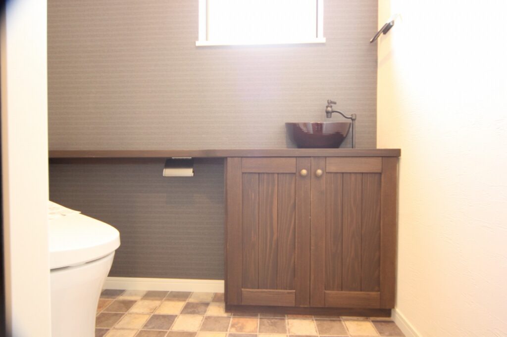 ダークグレーのアクセントクロスにクラシックな木製手洗いキャビネットとボウルを合わせたトイレ空間の画像