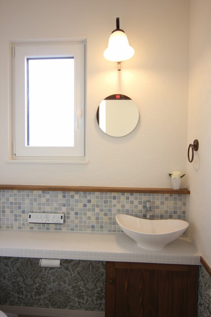 ブルーのモザイクタイルカウンターに白い手洗いボウルと丸型ミラーを合わせたエレガントなトイレの画像