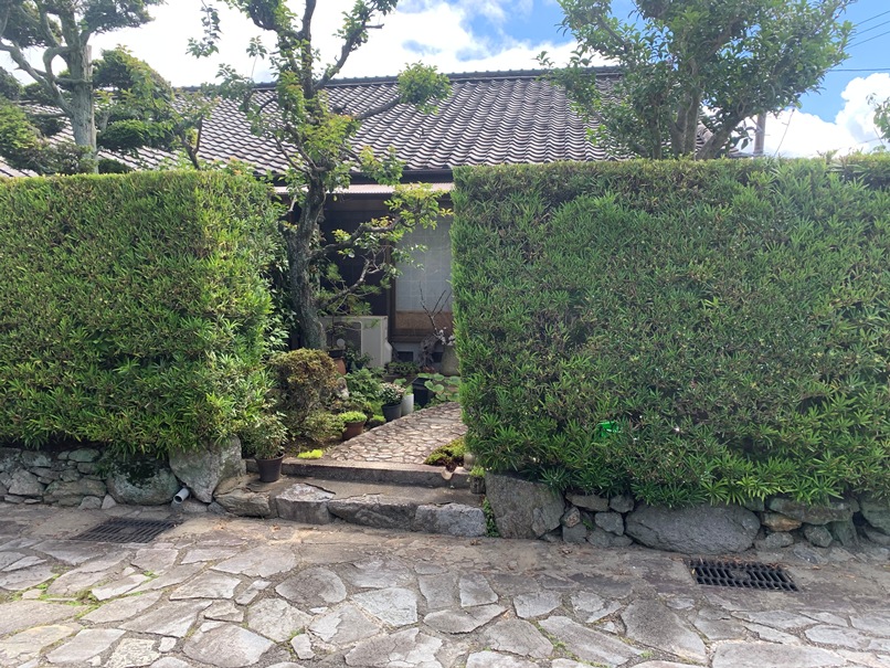 生垣の切れ目から前庭が覗く。鉢植えや室外機——「暮らし」がそこにあるの画像